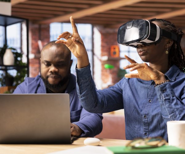 Black girl engages with virtual reality while her partner works on a laptop at the desk, exploring AI and interactive technology in a modern home office setting. 3D simulation with gadgets.